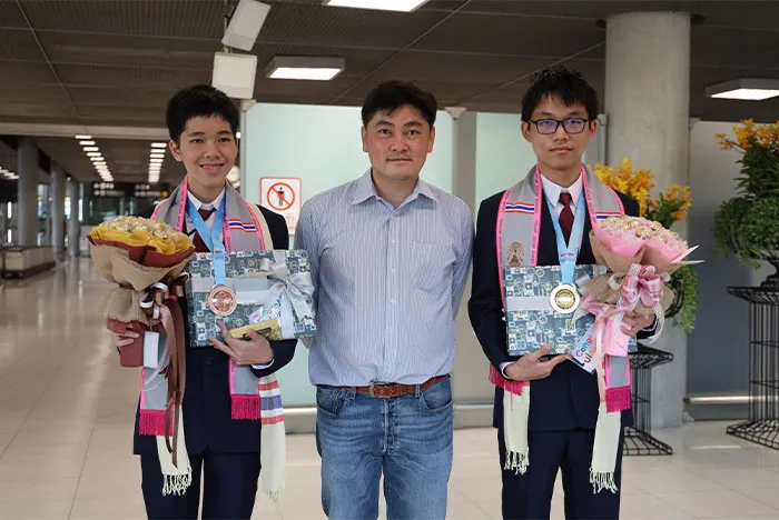 ภาพผู้สอนกับนักเรียนสองคน สวมเหรียญและถือช่อดอกไม้
